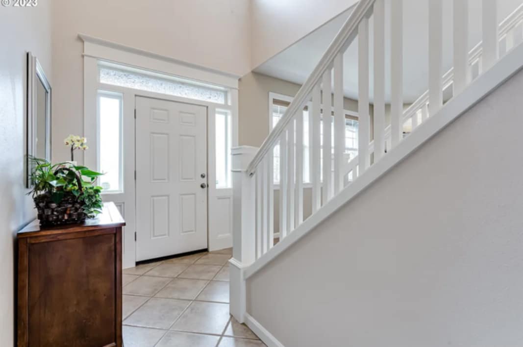 Photo of the front door of a house from the inside, a partial view of a staircase to the right and a cabinet with a plant on top to the left.